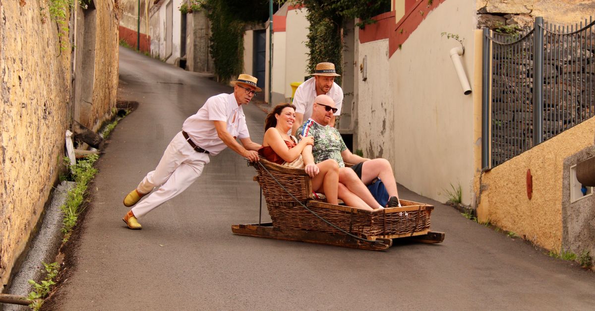 A wicker basket on wooden runners speeds down a steep street in Funchal, steered from behind by two men in white clothing with straw hats