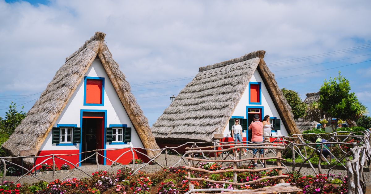 A whitewashed cottage with a steep, ground-length thatched roof and a red door, surrounded by planted flower beds