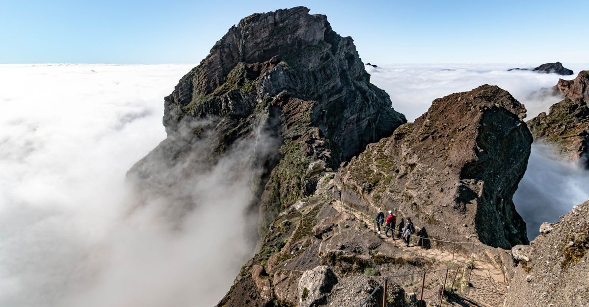 Two hikers on a narrow rock path high above an unbroken sea of cloud, bare basalt ridges all around